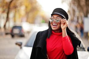 chica de moda afroamericana con abrigo, gorra de vendedor de periódicos y gafas de sol posadas en la calle contra un auto de negocios blanco. foto