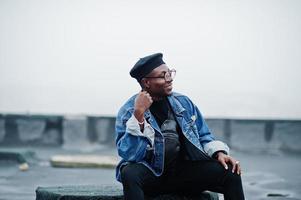 hombre afroamericano con chaqueta de jeans, boina y anteojos posados en un techo abandonado. foto