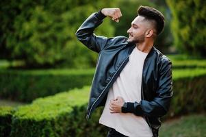 Stylish indian beard man at black leather jacket shows his muscle hand. India model posed at green garden of city. photo