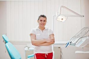 retrato de mujer dentista cruzó los brazos de pie en su oficina de odontología cerca de la silla. foto