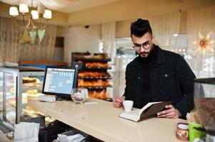 Arab man wear on black jeans jacket and eyeglasses in cafe drink coffee at bar with book. Stylish and fashionable arabian model guy. photo