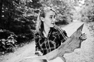 Portrait of a positive young gorgeous blonde sitting on the ground with a map in her hands in the forest. photo