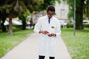 elegante médico afroamericano con estetoscopio y bata de laboratorio, con anteojos posados al aire libre y usando su teléfono móvil. foto