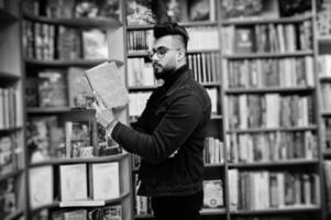 Tall smart arab student man, wear on black jeans jacket and eyeglasses, at library with book at hands. photo