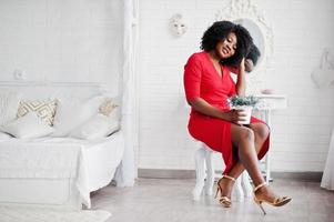 Fashion african american model in red beauty dress, sexy woman posing evening gown sitting at chair with pot of lavender against mirror in white vintage room. photo