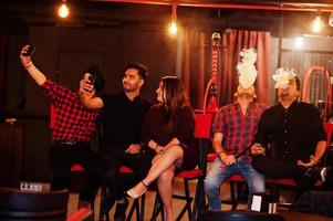 Group of indian friends sitting at lounge bar, having fun and rest, smoking hookah and looking on mobile phones to make selfie. photo