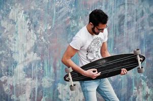 Street style arab man in eyeglasses with longboard posed against colored wall, like he play guitar. photo