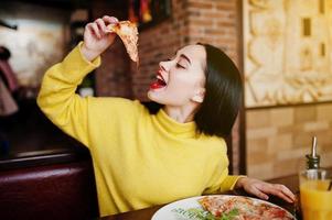 chica morena divertida en suéter amarillo comiendo pizza en el restaurante. foto