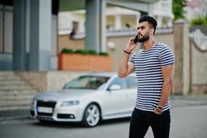 Handsome tall arabian beard man model at stripped shirt posed outdoor against car. Fashionable arab guy. Muslim businessman speaking at mobile phone. photo
