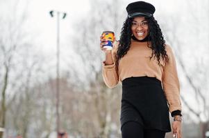 African american woman in black skirt, brown sweater and cap posed at winter day against snowy background, holding cup of coffee and phone. photo