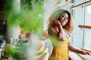 Glamour african american woman in yellow dress posed at restaurant near windows. photo