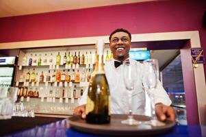 African american bartender at bar holding champagne with glasses on tray. photo