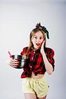 Young funny housewife in checkered shirt and yellow shorts pin up style with saucepan and kitchen spoon isolated on white background. photo