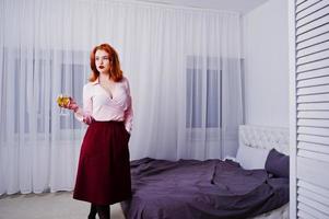 hermosa chica pelirroja con blusa rosa y falda roja con una copa de vino a mano cerca de la cama en la habitación. foto