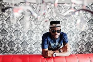 Rich african man in stylish traditional clothes and hat in glasses posed at room against vintage background, near red leather sofa bed. photo