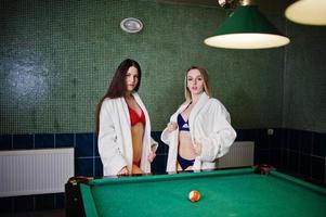 Two sexy girls in white bathrobe and swimsuit play pool billiards. photo