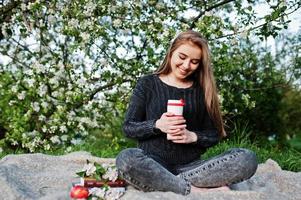 Young brunette girl at jeans sitting on plaid against spring blossom tree and holding tea thermos at hands. photo