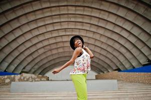 Amazing african american model woman in green pants and black hat posed outdoor against arena hall. photo