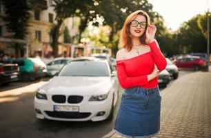 Attractive redhaired woman in eyeglasses, wear on red blouse and jeans skirt posing at street against white sport car. photo