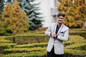 hombre indio elegante con bindi en la frente y anteojos, vestido con traje posado al aire libre contra arbustos verdes en el parque y mirando sus relojes. foto