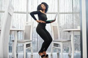 Fashionable young beautiful african american business woman with afro hairstyle wear in elegant black, stand with laptop at hands. photo