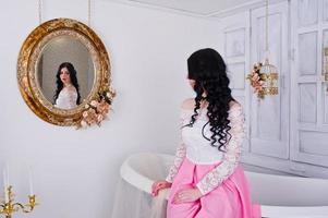 jovencita morena con falda rosa y blusa blanca posada en el interior de la habitación con baño decorativo y espejo. foto