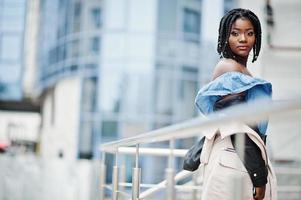 Attractive african american woman with dreads in jacket posed near railings against modern multistory building. photo