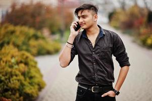 hombre indio con camisa marrón posó al aire libre y hablando por teléfono móvil. foto