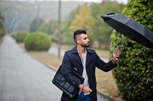 hombre de barba árabe alto de moda vestido con abrigo negro con paraguas y estuche posado en el día de la lluvia. foto