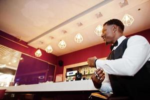 Young handsome african man wearing white shirt, black vest and bow tie with wallet purse posed against bar counter at night club. photo