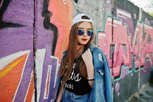 Stylish casual hipster girl in cap, sunglasses and jeans wear, listening music from headphones of mobile phone against large graffiti wall.