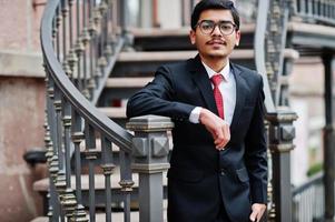 joven indio con anteojos, traje negro con corbata roja posado al aire libre contra escaleras de hierro. foto