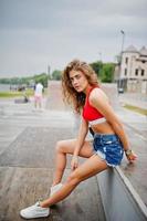 chica sexy modelo rizada en top rojo, pantalones cortos de mezclilla, anteojos y zapatillas posadas en skatepark. foto