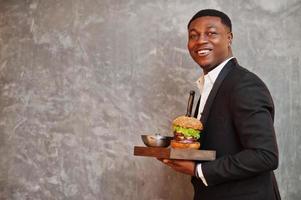 Respectable young african american man in black suit hold tray with double burger against gray wall. photo