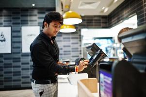 Indian man holding pay by credit card at cash desk with order screen and payment terminal in food cafe. photo