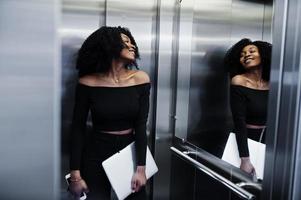 moda joven y hermosa mujer de negocios afroamericana con peinado afro en elegante negro, de pie en el ascensor con el teléfono y la computadora portátil en las manos. foto