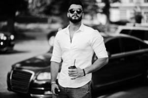 Stylish tall arabian man model in white shirt, jeans and sunglasses posed at street of city. Beard attractive arab guy with cup of coffee against black business car. photo