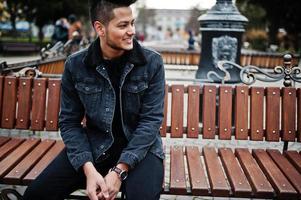 hombre indio guapo y de moda en chaqueta de jeans negros posó al aire libre, sentado en un banco. foto
