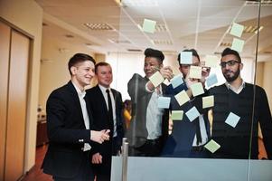 hombre de negocios apuntando sobre vidrio con notas de papel de colores. grupo diverso de empleados varones en ropa formal usando pegatinas. foto