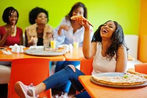 African woman with pizza sitting at restaurant against dark skinned girls. photo