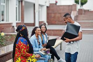 Group of five african college students spending time together on campus at university yard. Black afro friends studying at bench with school items, laptops notebooks. photo