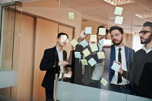 Business mans pointing on glass with colorful paper notes. Diverse group of male employees in formal wear using stickers. photo