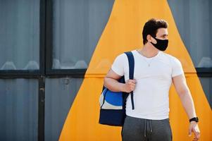 Portrait sports arabian man in black medical face mask with backpack posed against yellow triangle during coronavirus quarantine. photo
