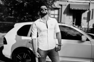 Stylish tall arabian man model in white shirt, jeans and sunglasses posed at street of city. Beard rich attractive arab guy against white suv car. photo