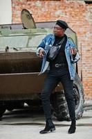 African american man in jeans jacket, beret and eyeglasses, with cigar posed against btr military armored vehicle. photo