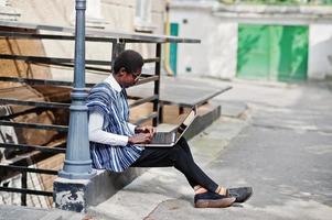 hombre africano con ropa tradicional y gafas con laptop trabajando al aire libre. foto