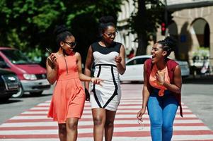 Three stylish african american womans walking on crosswalk or pedestrian crossing, speaking each other and having fun. photo