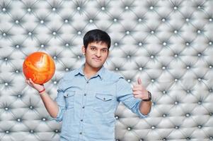 Stylish asian man in jeans shirt standing with bowling ball at hand against silver wall background and show thumb up. photo