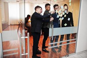 Business mans pointing on glass with colorful paper notes. Diverse group of male employees in formal wear using stickers. photo