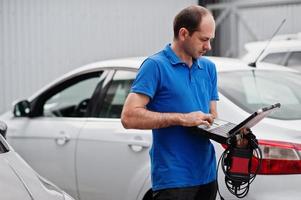 Car repair and maintenance theme. Electric mechanic in uniform working in auto service, making car diagnostics using obd device with laptop. photo